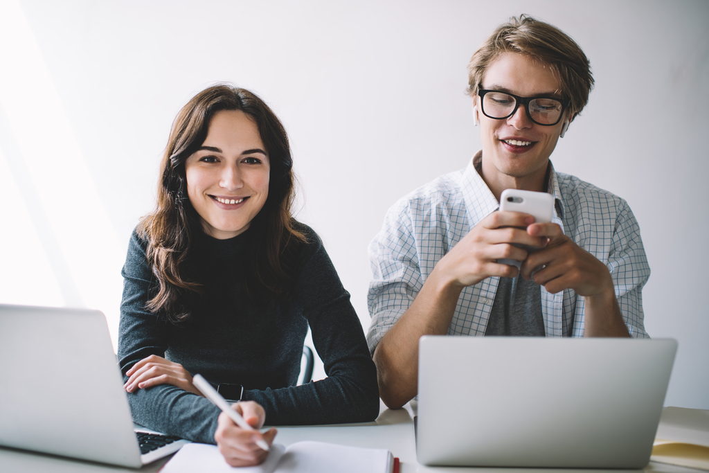 Portrait of cheerful caucasian woman millennial freelancer looking at camera doing homework with positive man blogger friend sending messages via mobile phone, hipster guy using app on cellular dwie młode osoby